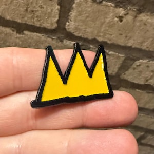 Könnte beinhalten: Ein gelber Kronen-Anstecker mit schwarzem Umriss. Der Anstecker hat ein einfaches, stilisiertes Design mit drei Spitzen. Die Krone wird in der Hand einer Person vor einem Ziegelhintergrund gehalten.