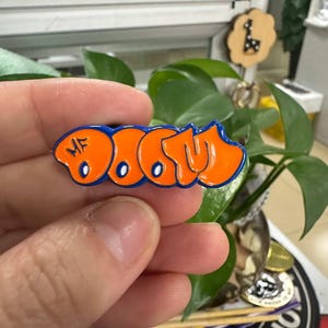 Op de afbeelding: Een oranje en blauwe emaille pin met het woord "DOOM" in een graffiti-stijl. De pin wordt in een hand gehouden voor een groene plant. De pin is een klein accessoire.