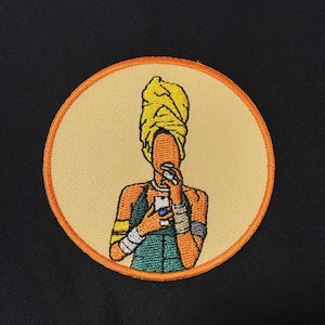 Peut inclure: Écusson brodé représentant une illustration stylisée d'une personne portant un turban jaune, tenant un téléphone et une tasse. L'écusson a une bordure orange et un fond beige, sur fond noir.
