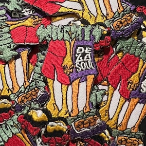 May include: A collection of embroidered patches with a colorful, cartoonish design. The patches feature figures with red, purple, yellow, and green accents, and the text "DE ZA SOUL". The patches are on a black background.
