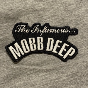 Op de afbeelding: Zwart en wit geborduurde patch met de tekst "The Infamous... MOBB DEEP". De patch heeft een zwarte achtergrond met witte letters en een witte rand. De patch ligt op een grijze stof.