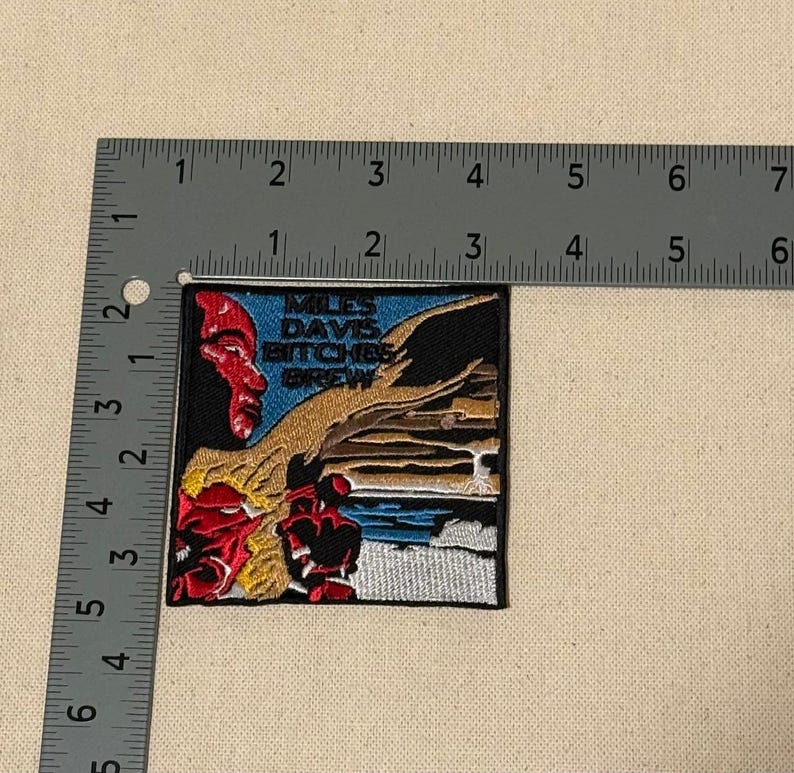 May include: Square embroidered patch featuring artwork from the Miles Davis album "Bitches Brew." The design includes a red and blue portrait, the album title, and abstract imagery. The patch measures approximately 2.5 inches by 2.5 inches.