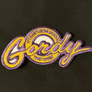 Puede incluir: Parche bordado con el nombre "Gordy" en escritura amarilla estilizada, contorneado en morado. El parche incluye el texto "IT'S WHAT'S IN THE GROOVES THAT COUNT" en un diseño circular. El parche está sobre un fondo negro.