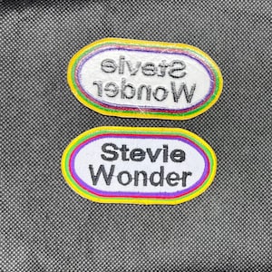 May include: Two embroidered patches with the text "Stevie Wonder" in a white oval with a colorful border. One patch is right-side up, and the other is upside down. The patches are on a textured gray surface.