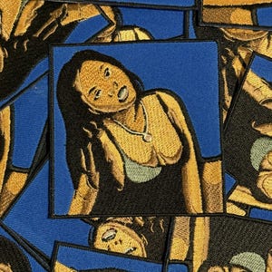 Könnte beinhalten: Gestickter Aufnäher mit einer stilisierten Darstellung einer Person mit langen dunklen Haaren, die eine Halskette und ein Oberteil trägt. Der Aufnäher hat einen blauen Hintergrund und goldene und schwarze Details. Mehrere Aufnäher sind im Bild zu sehen.