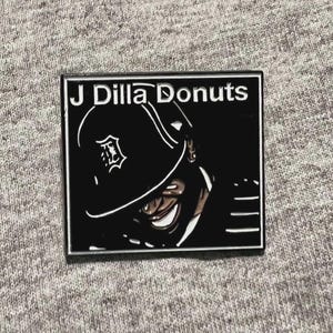 May include: A rectangular black and white enamel pin featuring an illustration of a person wearing a baseball cap. The pin has the text "J Dilla Donuts" in white lettering.