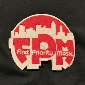 May include: A red and white embroidered patch with the text "First Priority Music" and the letters "FPM" in a stylized font. The patch features a cityscape silhouette at the top and is set against a black background.