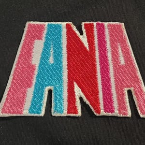 Pode incluir: Patch bordado com o nome "ANA" em letras maiúsculas. As letras são rosa, azul e vermelho, com contornos brancos, sobre um fundo preto. Ideal para personalizar roupas ou acessórios.