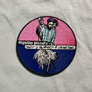 Könnte beinhalten: Gestickter Aufnäher mit dem Text "digable planets" und "reachin' (a new refraction of time and space)". Der Aufnäher hat ein rosa-blaues Farbschema mit einer Illustration von drei Personen und einem pflanzenartigen Design.