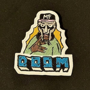 Pode incluir: Patch bordado com um personagem mascarado com o texto "MF" em um chapéu. O patch tem uma borda branca e a palavra "DOOM" em letras azuis.