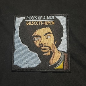 May include: Embroidered patch featuring a portrait of Gil Scott-Heron with the text "PIECES OF A MAN" above his image. The patch has a black border, a blue background, and is set against a black fabric.