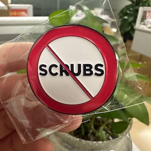 Può includere: Una spilla rotonda con bordo rosso e centro bianco. La parola "SCRUBS" è stampata in nero, con una linea diagonale rossa che la attraversa. La spilla è in un involucro di plastica trasparente.