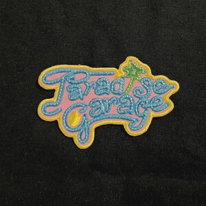 May include: Embroidered patch with the words "Paradise Garage" in a stylized script. The patch features a pink background with blue and yellow accents, and a small green palm tree graphic.