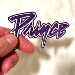 May include: Embroidered purple "Prince" name patch with a white border. The patch is held by a hand against a white background. The patch is a music-themed item, suitable for clothing or accessories.