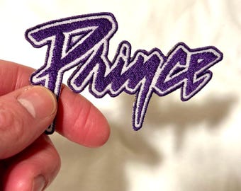 May include: Purple embroidered patch with the name "Prince" in a stylized script. The patch has a white border and is held by a hand against a white background. The patch is a music-themed item.