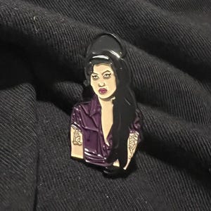 May include: Enamel pin featuring a portrait of a woman with long black hair, wearing a purple shirt. The pin has a glossy finish and depicts tattoos on the arms. The pin is set against a dark background.