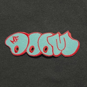 Può includere: Toppa ricamata con la parola "DOOM" in un carattere stilizzato. Le lettere sono azzurre con un contorno e accenti rossi. La toppa è su sfondo nero.