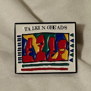 Puede incluir: Pin rectangular de esmalte con el nombre de la banda "TALKING HEADS" en blanco sobre un borde negro. El diseño del pin incluye figuras abstractas en rojo, amarillo, azul y verde. El fondo del pin es blanco.