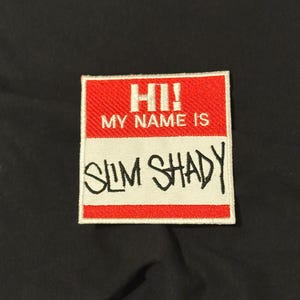 Puede incluir: Un parche bordado rectangular que presenta una sección superior roja con el texto blanco HI! MY NAME IS y una sección inferior blanca con el texto negro bordado SLIM SHADY. El parche tiene un borde blanco sobre un fondo de tela negra.