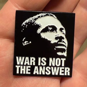 May include: Black square pin featuring a white portrait and the text "WAR IS NOT THE ANSWER". The pin is held in a hand.