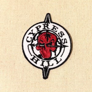 Peut inclure: Écusson brodé avec un crâne rouge au centre, entouré d'un cercle blanc avec le texte "CYPRESS HILL" en noir. Le patch comporte des flèches noires et une bordure noire, sur un fond en tissu beige.