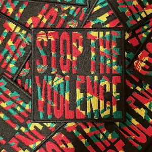 Può includere: Toppa quadrata nera con le parole "STOP THE VIOLENCE" ricamate in rosso, giallo e verde. La toppa ha un bordo nero ed è circondata da toppe simili.