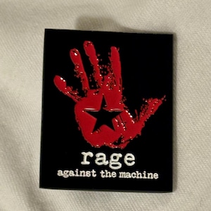 May include: A black rectangular pin badge with a red handprint and a black star in the centre. The words "rage against the machine" are printed in white below the handprint.