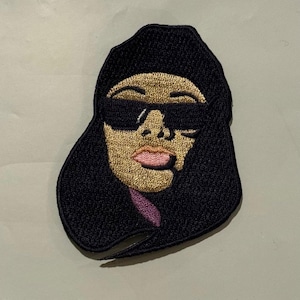 Puede incluir: Parche bordado con un rostro estilizado con gafas de sol y un gorro negro. La cara tiene piel bronceada, labios rosados y un cuello morado. El parche está contorneado en negro.