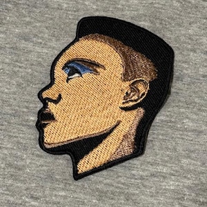 Könnte beinhalten: Gestickter Aufnäher mit einem Seitenprofil eines Gesichts. Der Aufnäher hat einen schwarzen Rand und zeigt das Gesicht in Braun- und Beigetönen mit blauem Lidschatten. Der Aufnäher befindet sich auf einem grauen Stoffhintergrund.
