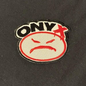 May include: Embroidered patch with the word "ONY" in black letters outlined in white, above a red cross. The patch features a white oval with a red outline and a red, frowning mouth.