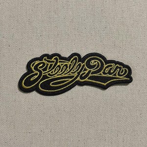 May include: A black embroidered patch with the words "Steely Dan" in a gold, cursive font. The patch has a black border and is set against a neutral fabric background. This patch is suitable for clothing or accessories.