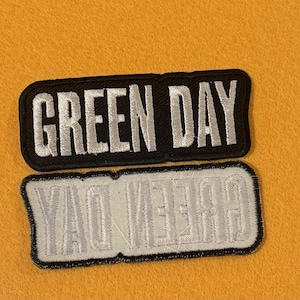 Könnte beinhalten: Zwei rechteckige Aufnäher mit dem Text "GREEN DAY". Der obere Aufnäher hat einen schwarzen Hintergrund mit weißer gestickter Schrift. Der untere Aufnäher ist ein Spiegelbild des oberen Aufnähers, mit weißem Hintergrund und schwarzer Schrift.