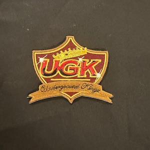 Peut inclure: Écusson brodé avec un motif de bouclier marron, avec le texte "UGK" en rouge et noir, surmonté d'une couronne dorée. En dessous, les mots "Underground Kings" sont brodés en or. L'écusson est posé sur un fond noir.