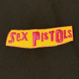 May include: A yellow embroidered patch with the words "Sex Pistols" in pink lettering. The patch has a pink border and is set against a black background. The patch is rectangular with slightly curved edges.