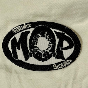 Puede incluir: Un parche bordado con las palabras "Firing MOP Squad" en letras blancas sobre un óvalo negro. El parche presenta un diseño negro estilizado en el centro. El fondo es una tela de color crema.