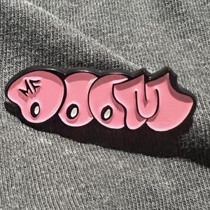Op de afbeelding: Een roze emaille pin met het woord "DOOM" in graffiti-stijl, omlijnd met zwart. De pin heeft de initialen "MF". De pin ligt op een grijze stoffen achtergrond.