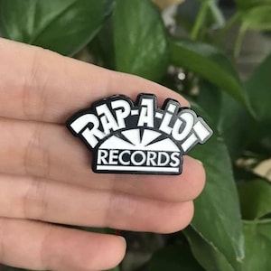 Peut inclure: Épinglette émaillée noire et blanche avec le texte "RAP-A-LOT RECORDS". Le design de l'épinglette est stylisé avec un élément en forme d'éclat d'étoile. L'épinglette est tenue dans une main devant un fond de feuilles vertes.