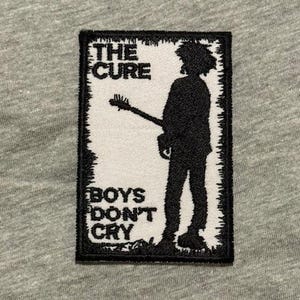 May include: A rectangular patch with a black border and the words "THE CURE" above "BOYS DON'T CRY". The patch features a black silhouette of a person playing a guitar on a white background. The patch is on a gray fabric.