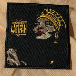 Könnte beinhalten: Schwarzer, gestickter Aufnäher mit dem Profil einer Person, die eine goldene Kopfbedeckung und roten Lippenstift trägt. Der Aufnäher enthält den Text "Queen Latifah" in goldener und roter Schrift.