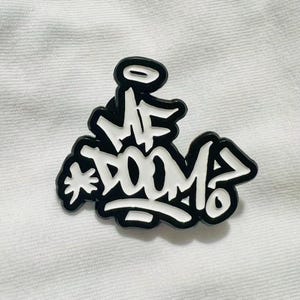 以下が含まれることがあります： 黒と白のピンバッジ。グラフィティ風のフォントで「MF DOOM」と書かれています。ピンバッジは黒い縁取りがあり、白い背景に配置されています。大胆で人目を引くデザインです。