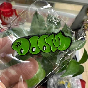 Op de afbeelding: Een groene emaille pin met het woord "DOOM" in graffiti-stijl, omlijnd in zwart. De pin zit in een doorzichtige plastic zak, met de letters "MF" zichtbaar.