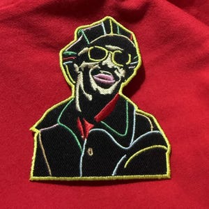 May include: Embroidered patch of a smiling man in a hat and sunglasses. The patch has a black coat, a red bow tie, and a yellow outline. The background is a solid red colour.