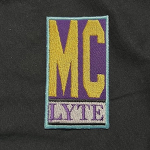 Könnte beinhalten: Gestickter Aufnäher mit den Buchstaben "MC" in Gold auf lila Hintergrund. Das Wort "LYTE" steht darunter in Grau. Der Aufnäher hat einen türkisfarbenen Rand und befindet sich auf schwarzem Stoff.