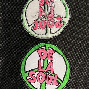 Könnte beinhalten: Zwei gestickte Aufnäher mit dem Schriftzug "De La Soul" in Rosa, innerhalb eines weißen Friedenszeichens mit grünem Rand. Die Aufnäher befinden sich auf schwarzem Hintergrund.