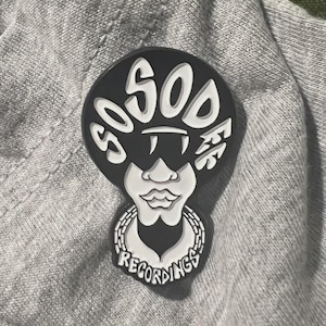 以下が含まれることがあります： スタイリッシュなフォントで「SOSOS」の文字が書かれた白黒のピンバッジ。サングラスをかけた顔と、下部に「RECORDINGS」の文字が描かれています。ピンバッジは灰色の布の背景に配置されています。