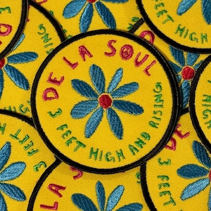 Könnte beinhalten: Gelbe runde Aufnäher mit blauem Blumenmuster und dem Text "DE LA SOUL" in Rosa. Der Aufnäher hat auch den Text "3 FEET HIGH AND RISING" in Grün. Die Blume hat eine rote Mitte und der Aufnäher hat einen schwarzen Rand.