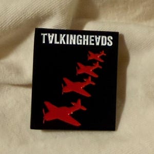 May include: Black square pin with the band name "TALKINGHEADS" in white. Red airplane silhouettes are arranged diagonally. A collectible item or accessory, possibly for a jacket or bag.