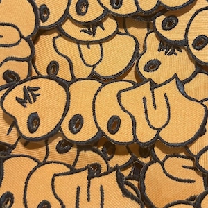 Peut inclure: Gros plan sur de nombreux écussons en tissu jaune avec des contours brodés noirs. Chaque écusson présente un motif stylisé avec les lettres "MF" et un détail circulaire. Les écussons sont densément tassés, créant une surface texturée.