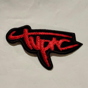 Puede incluir: Parche bordado negro con el nombre "Tupac" en escritura roja. El parche tiene una forma única con una parte superior curva y un borde inferior puntiagudo. El texto está cosido en una fuente audaz y estilizada.