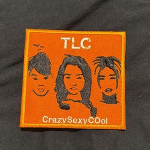 Può includere: Toppa ricamata arancione con tre volti stilizzati e la scritta "TLC" in alto. Sotto i volti, è visualizzato il testo "CrazySexyCool". La toppa ha un bordo giallo ed è posta su uno sfondo nero.